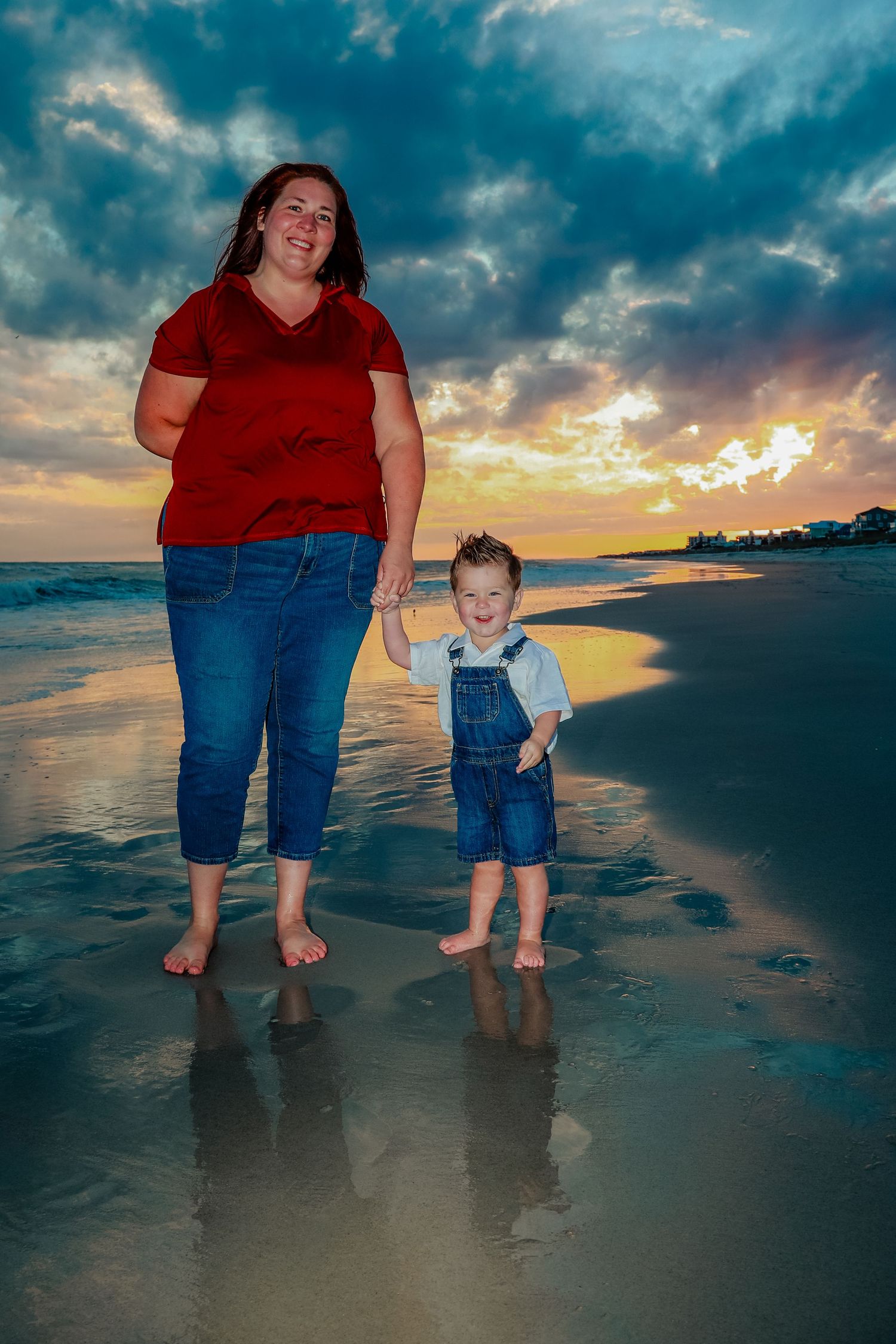 A mother and young child hold hands on a beach at sunset, both barefoot with reflections in the wet sand.