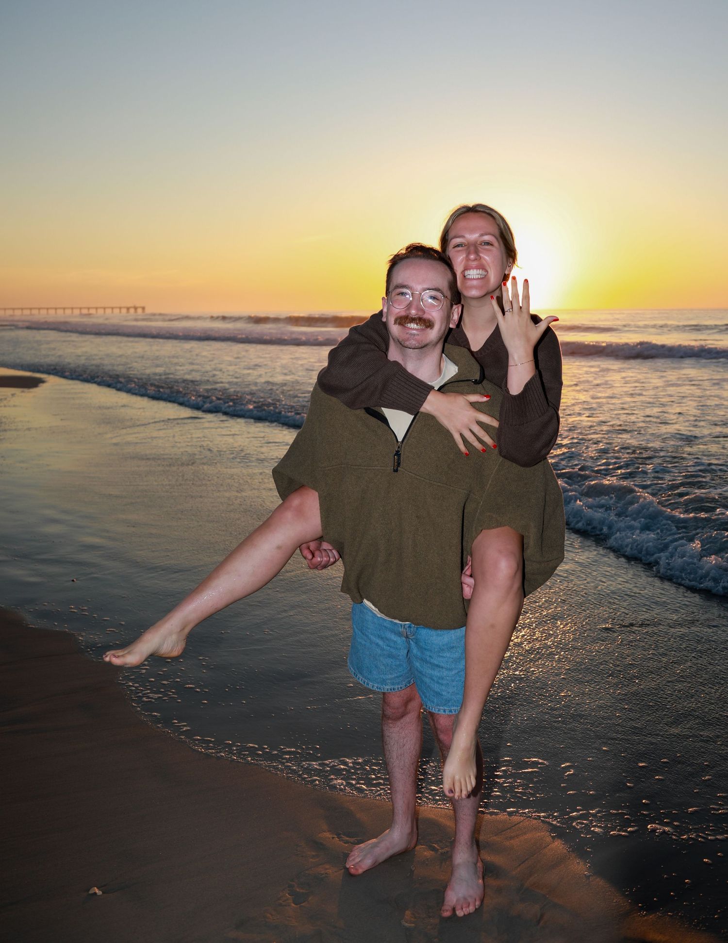 Couple celebrating engagement at sunset beach, woman showing ring while getting piggyback ride, ocean waves in background.