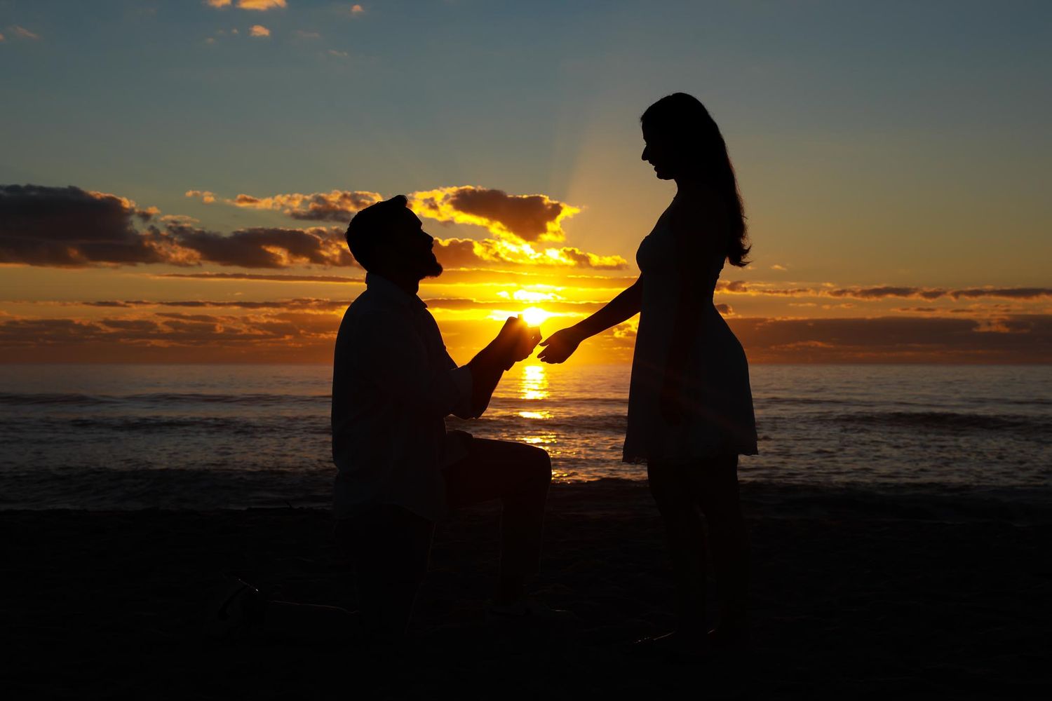 Silhouette of a man proposing on one knee to a woman on a beach at sunset.