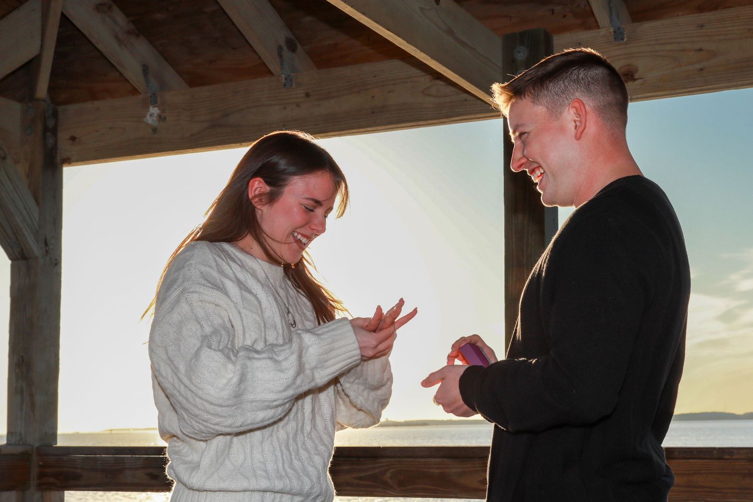 Couple sharing a romantic proposal moment at sunset on a wooden deck, man placing ring on womans finger.