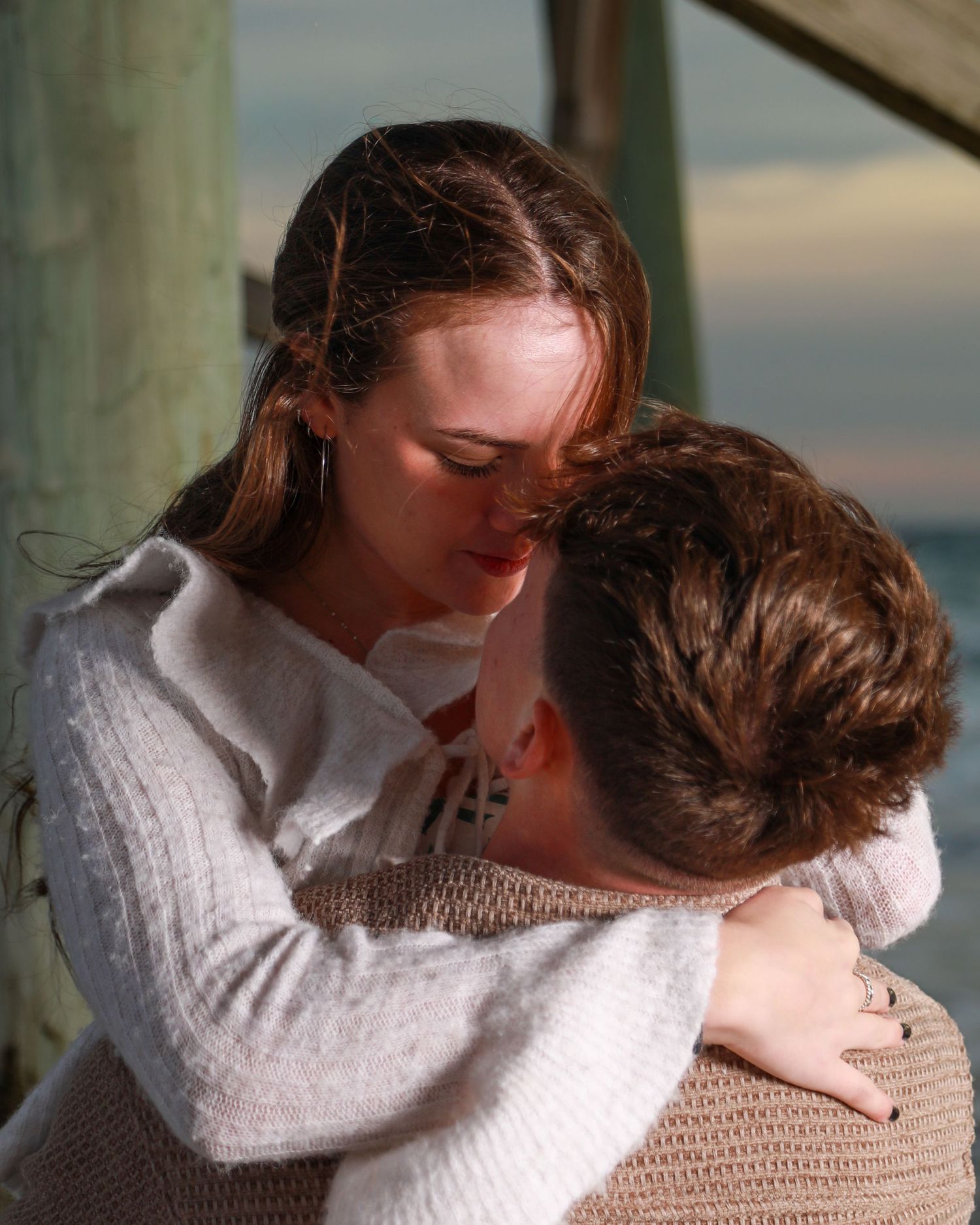 Two young people sharing a tender embrace outdoors near water at dusk, wearing cozy sweaters.