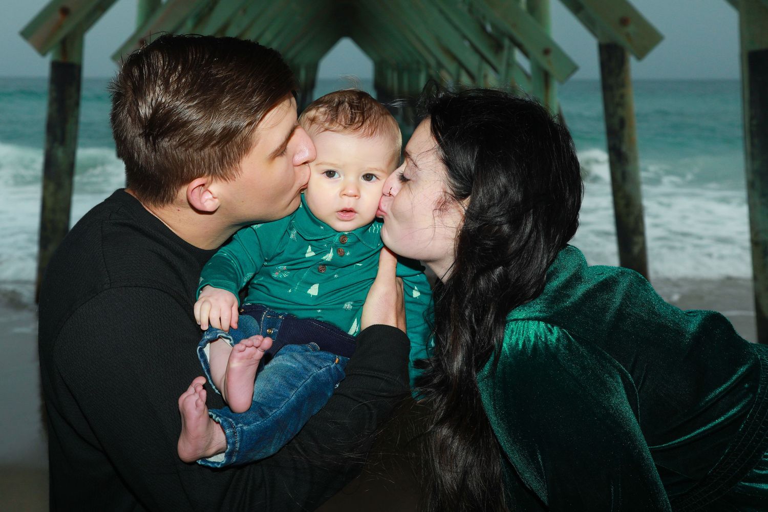 Two parents kissing their baby on the cheeks at a beach pier, all wearing green outfits.