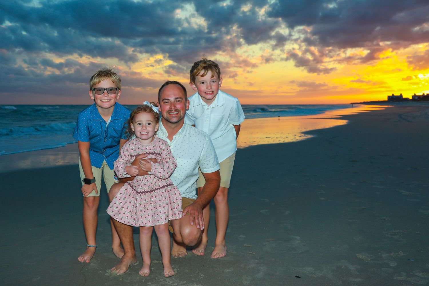 A father poses with three young children on a sandy beach during a vibrant sunset with colorful clouds.