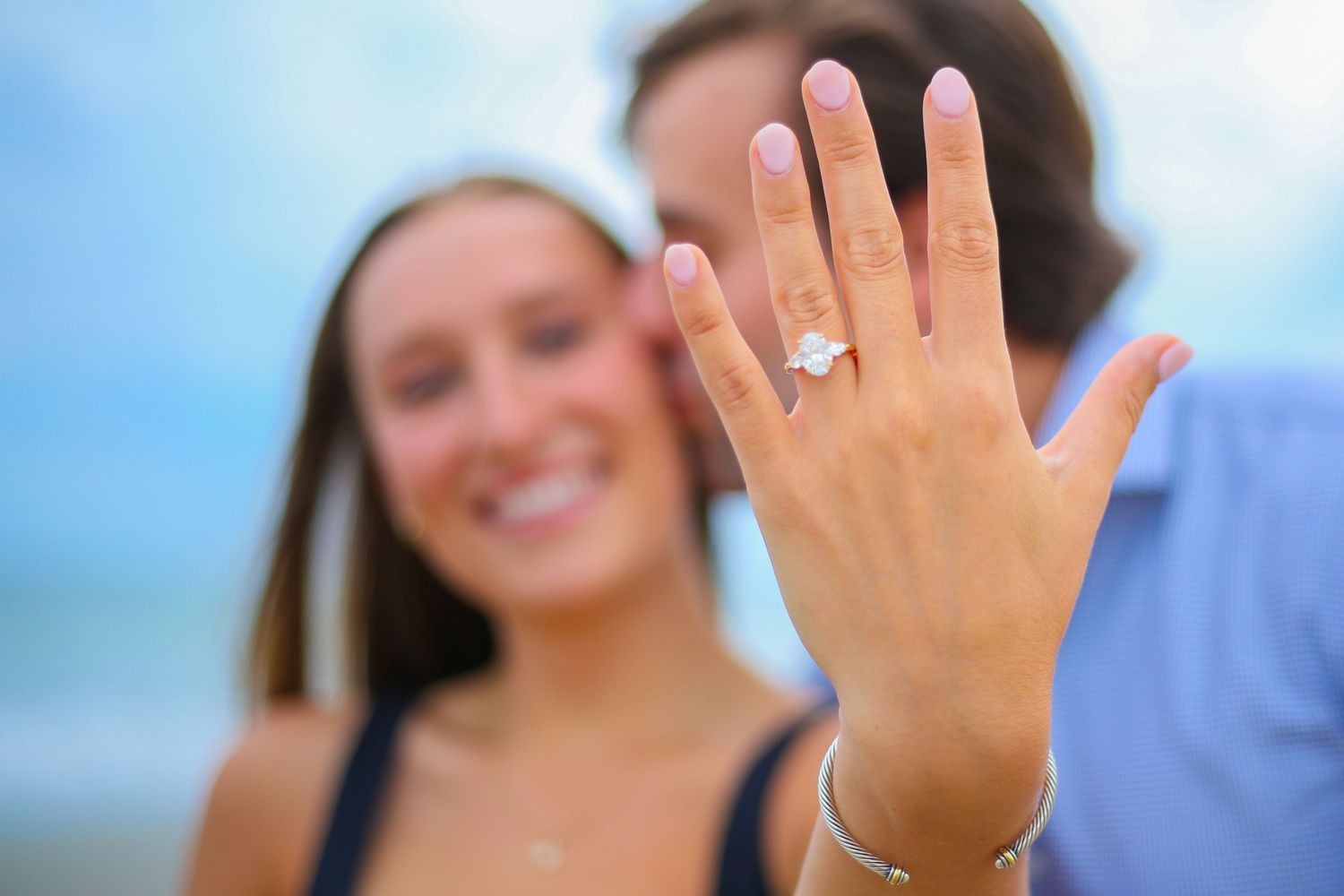 Smiling couple at beach showing off diamond engagement ring on woman's outstretched hand in sharp focus.