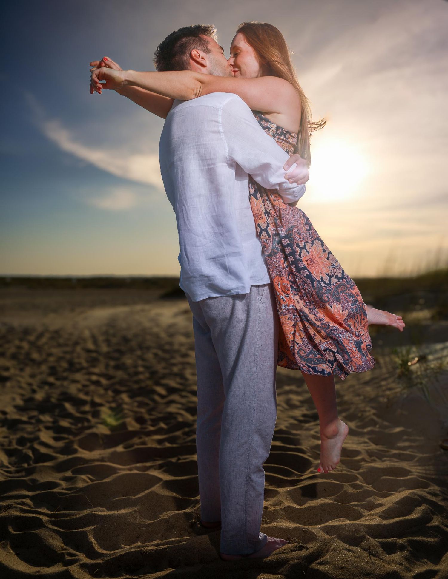 Romantic couple embracing and kissing on a sandy beach at sunset, man lifting woman in floral dress.