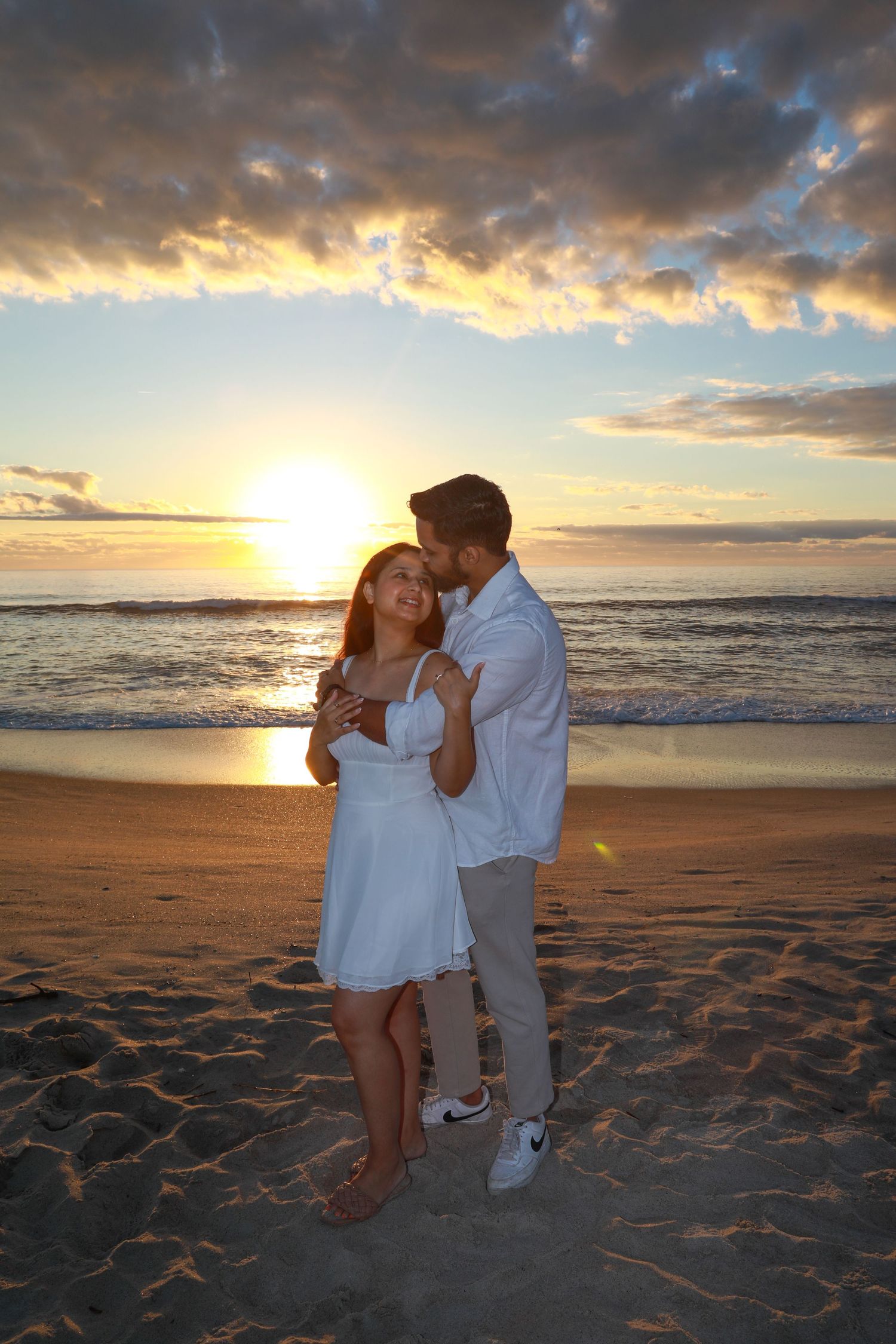Romantic couple embracing on a sandy beach at golden sunset, woman in white dress, waves in background.