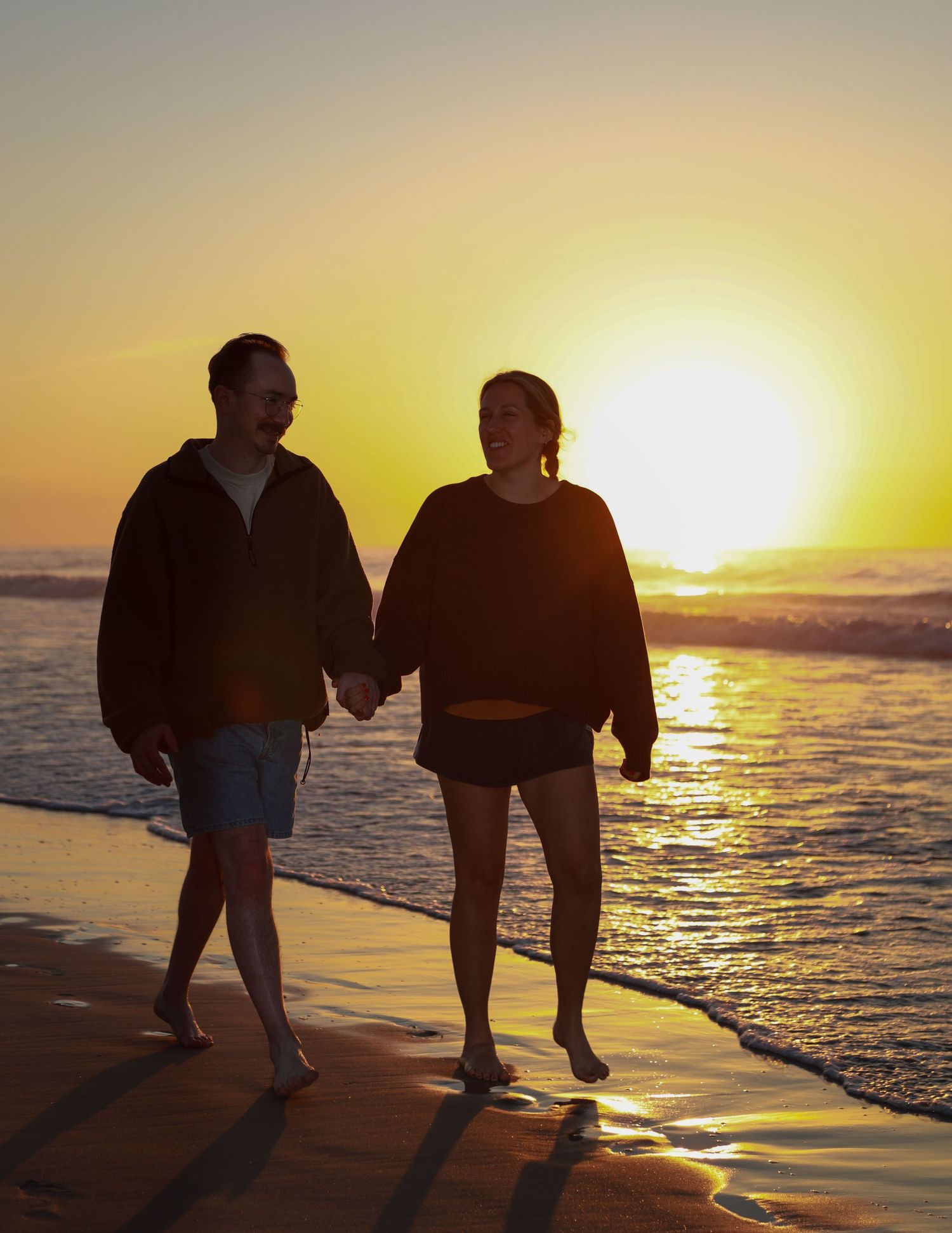 Couple holding hands walking barefoot on a sandy beach at golden sunset with ocean waves in the background.