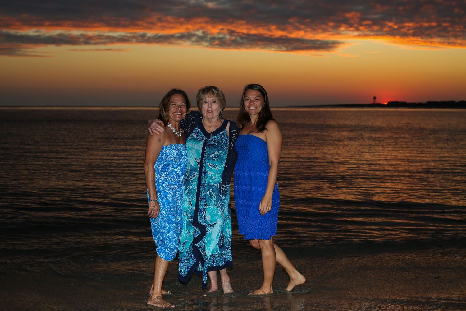 Three women pose smiling on a beach at sunset with vibrant orange sky and ocean waves behind them.