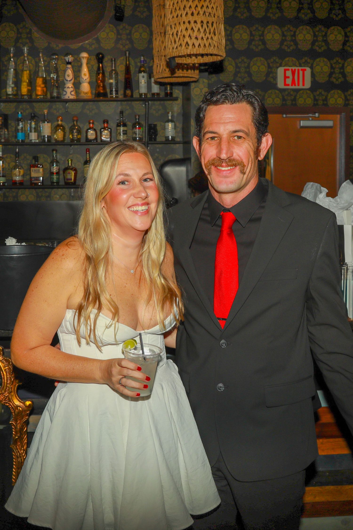 A smiling couple poses at a bar, woman in white dress holding a drink, man in dark suit with red tie.