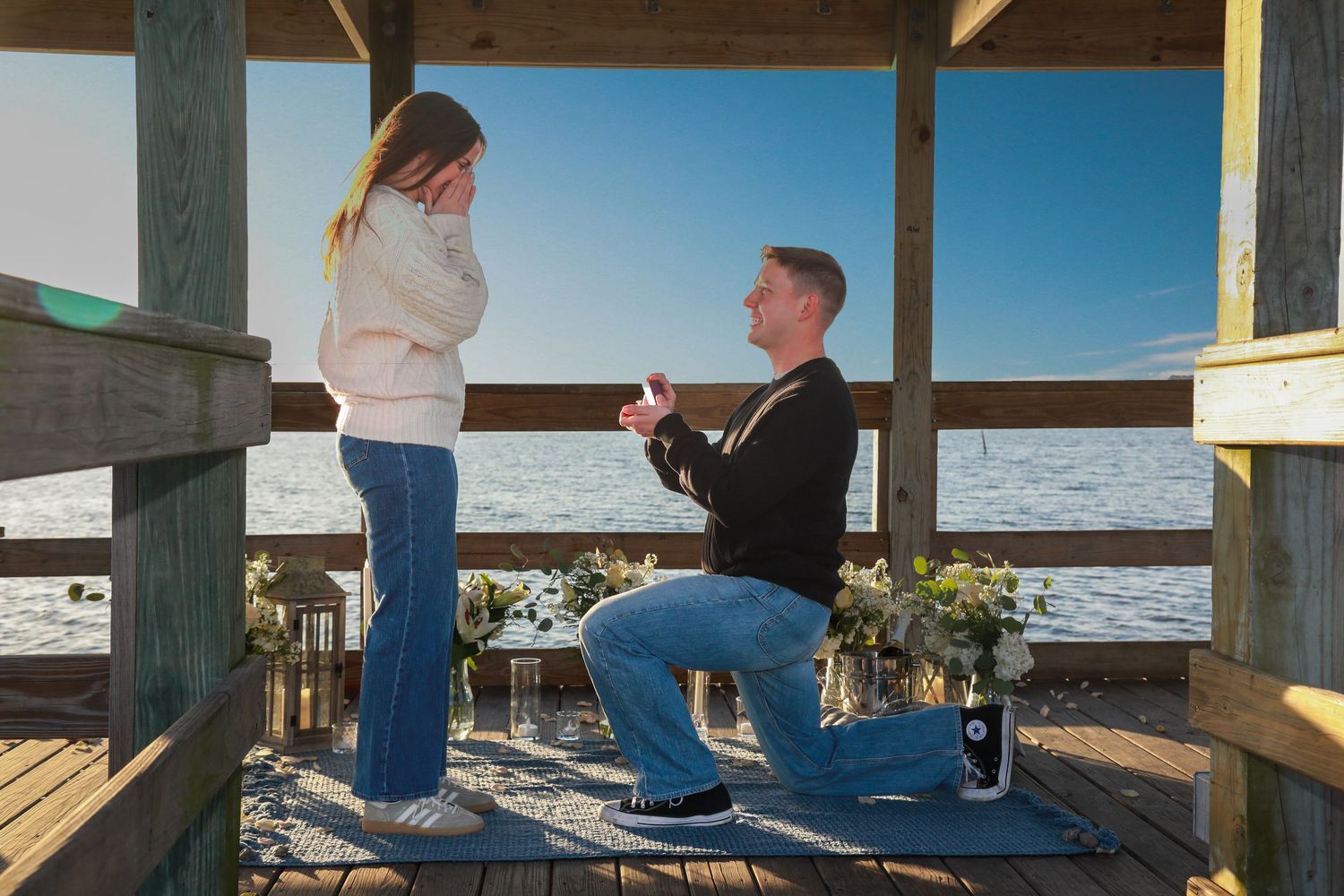 Man proposing to woman on a waterfront pier deck, holding a ring box while kneeling on one knee.