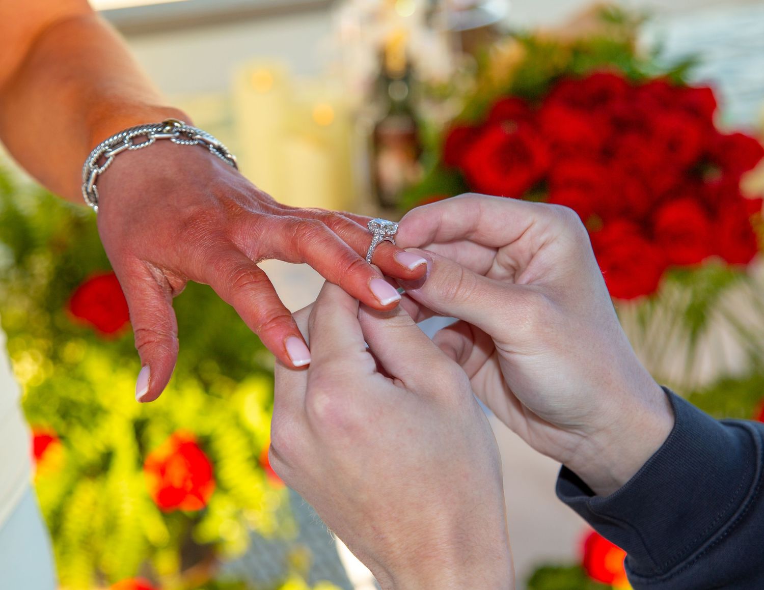 Person placing a diamond engagement ring on a woman's hand with red roses in the background.