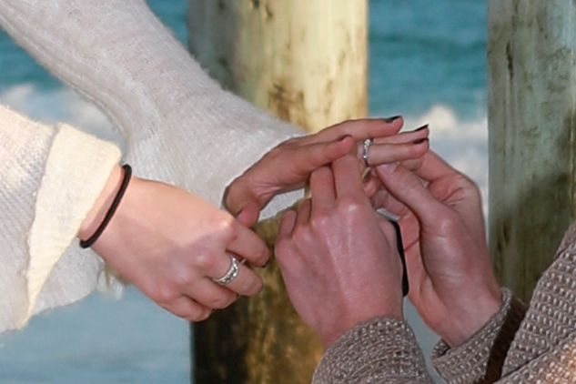 Two people exchanging rings near a pier by the ocean, symbolizing an engagement or wedding ceremony.