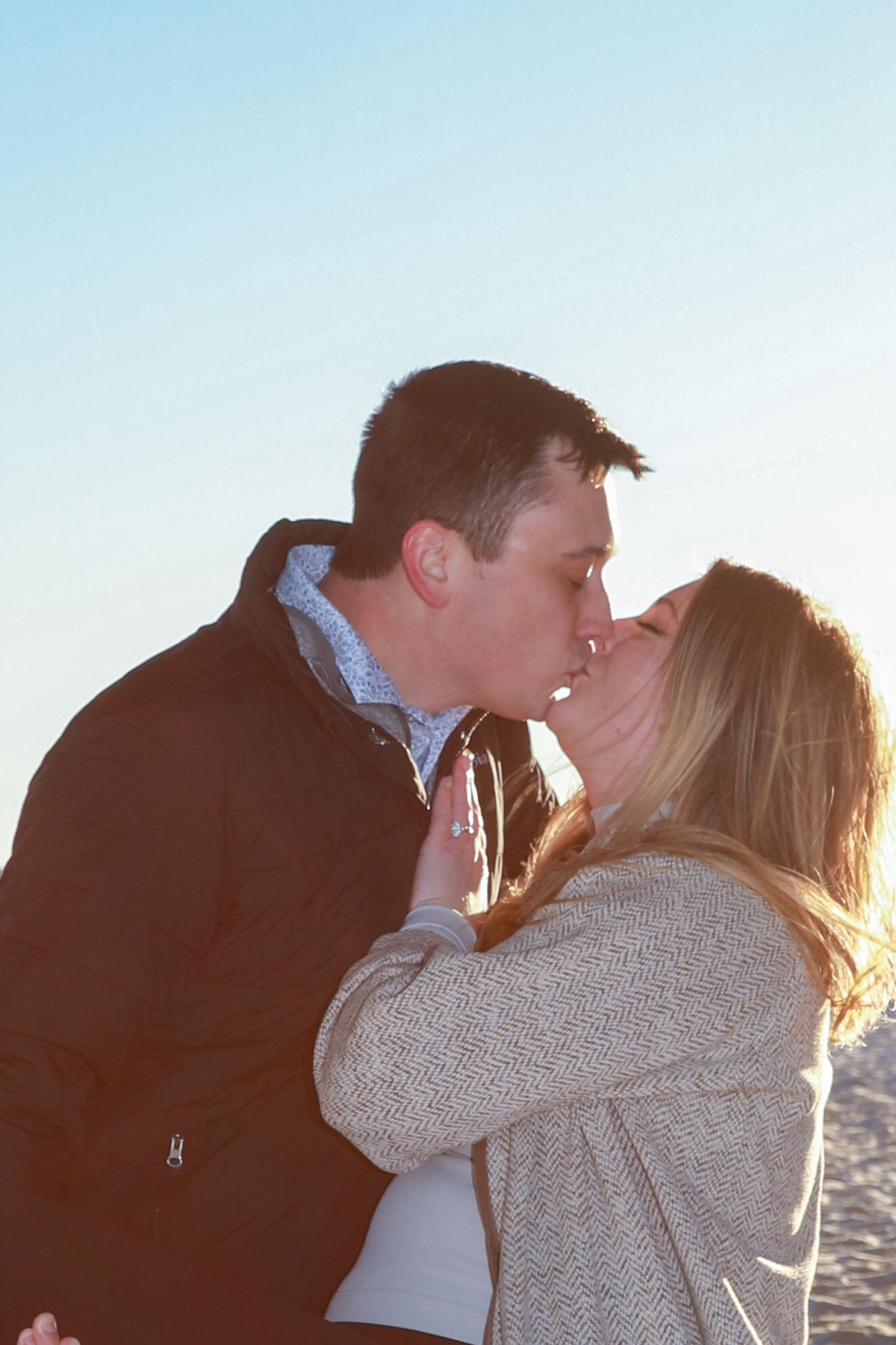 Couple sharing a romantic kiss outdoors on a sunny day, wearing casual jackets with a clear blue sky background.