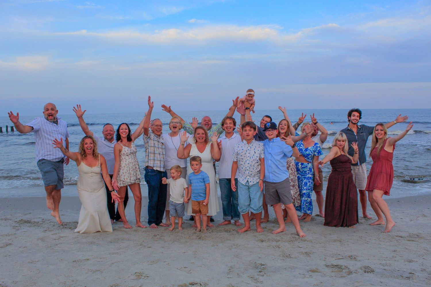Large family group posing joyfully on a sandy beach at sunset, waving hands in the air near ocean waves.