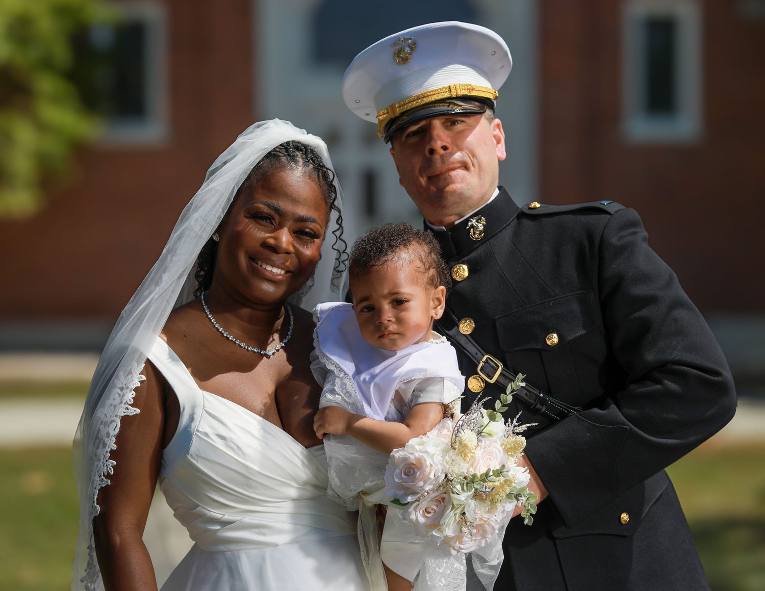 Military groom in dress uniform with bride in white wedding gown holding baby with bouquet outdoors.