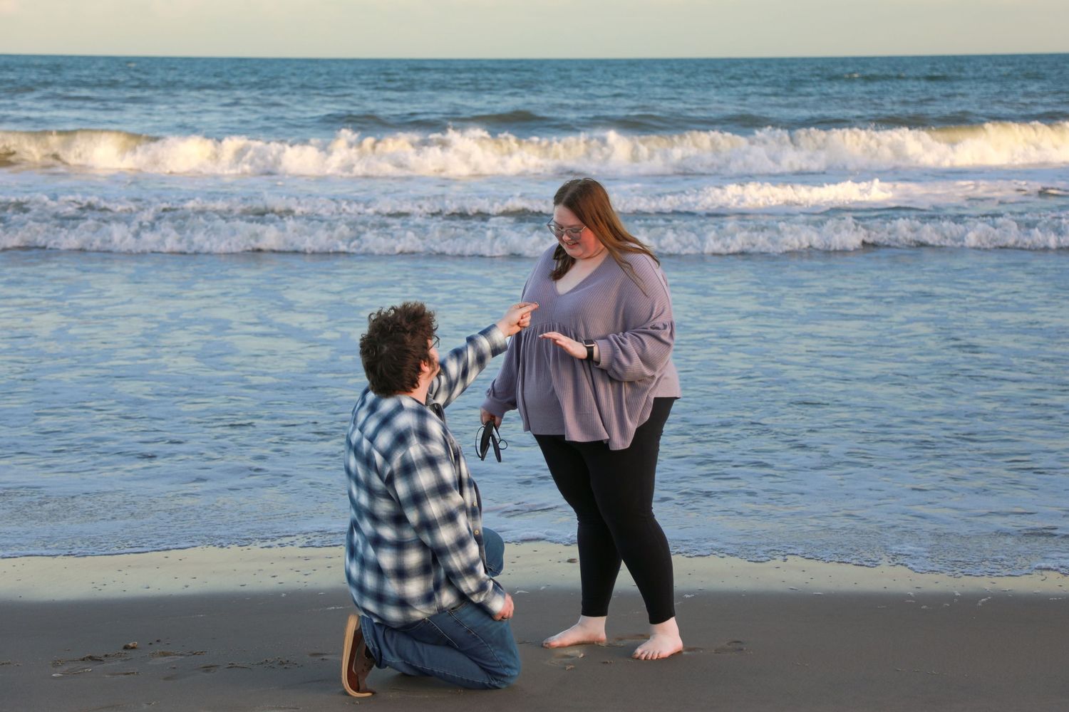 Man kneeling on beach proposing to woman at sunset with ocean waves in background.