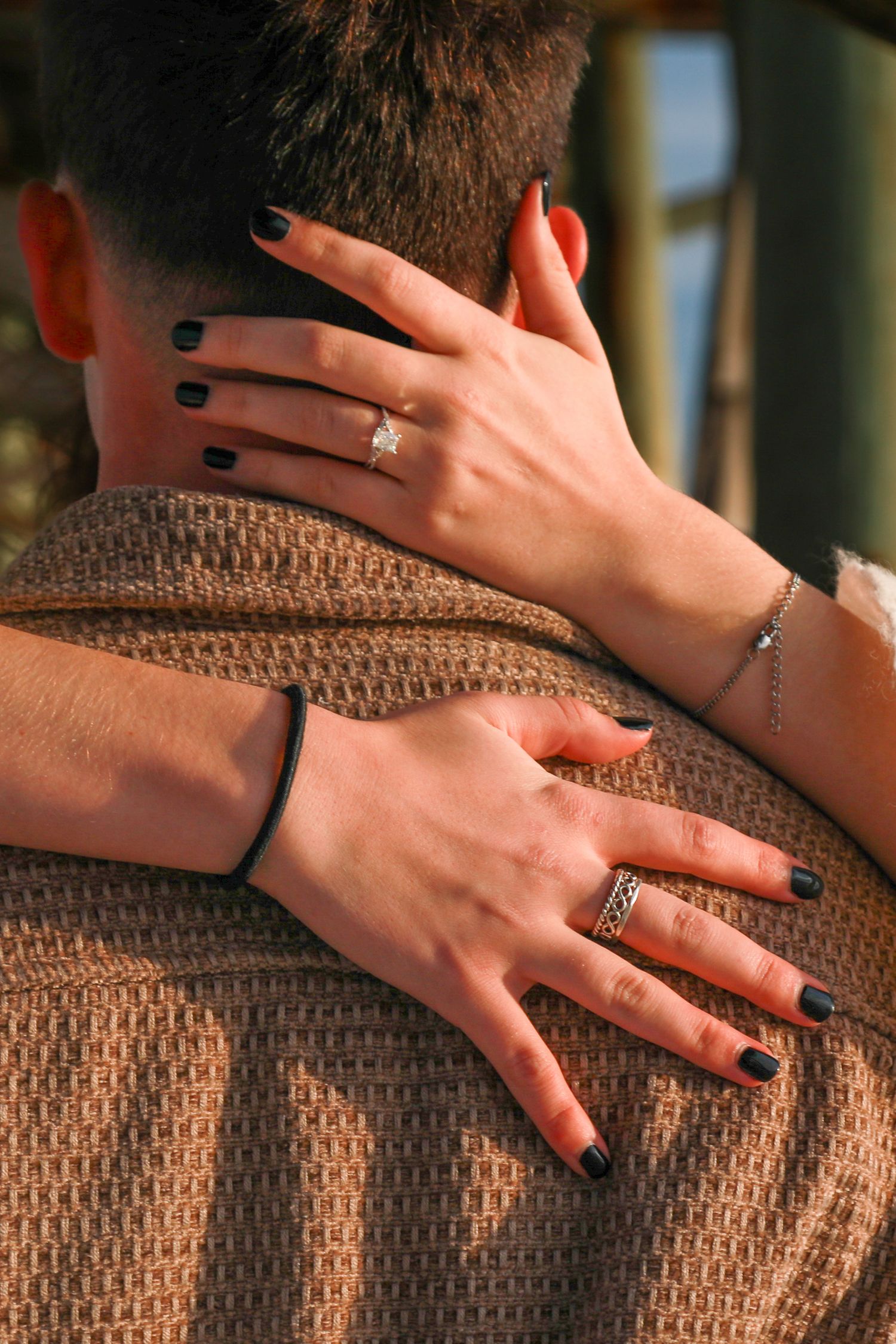 Woman with dark nails and diamond engagement ring embracing a man from behind, showcasing elegant jewelry.