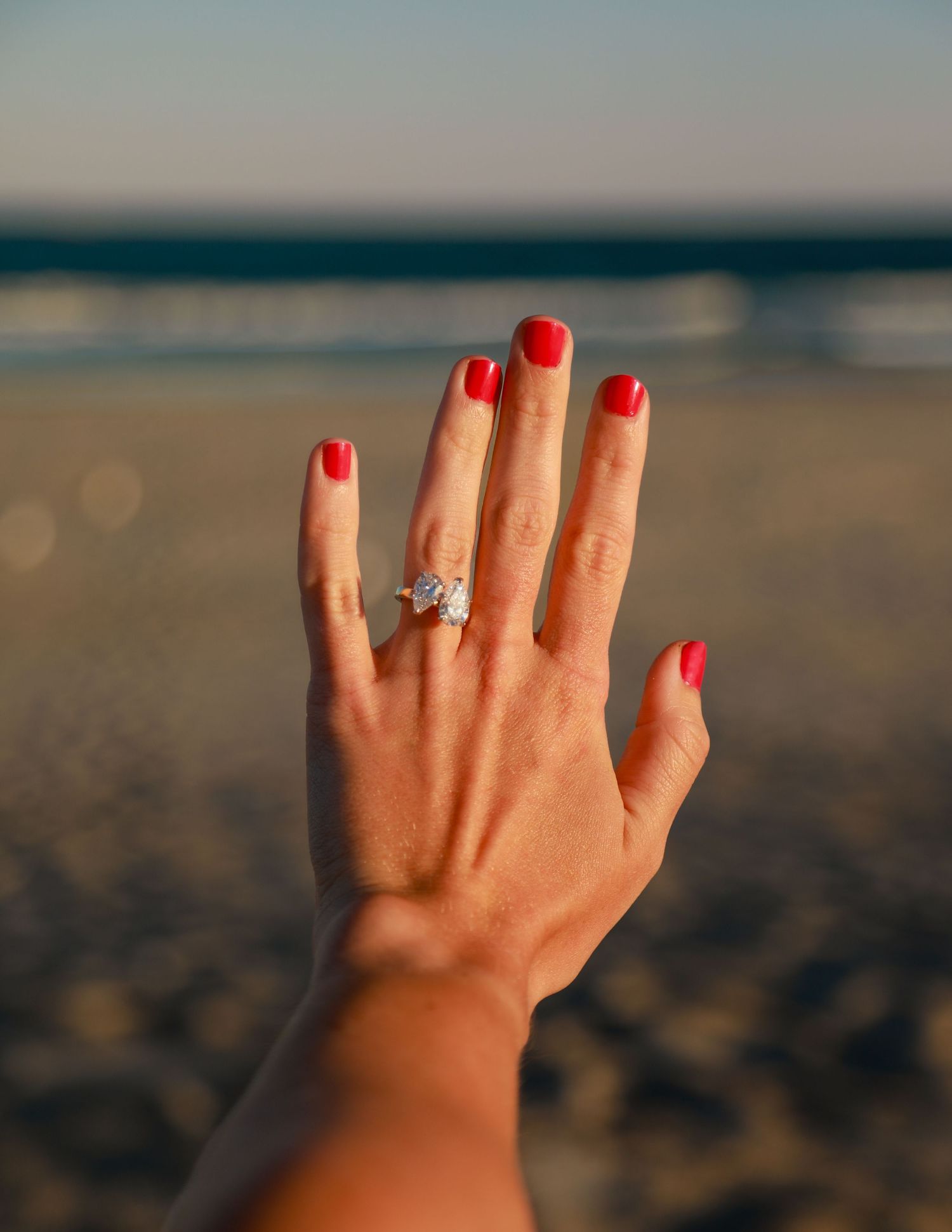 Close-up of a hand with red nails displaying a sparkling diamond engagement ring on a beach.