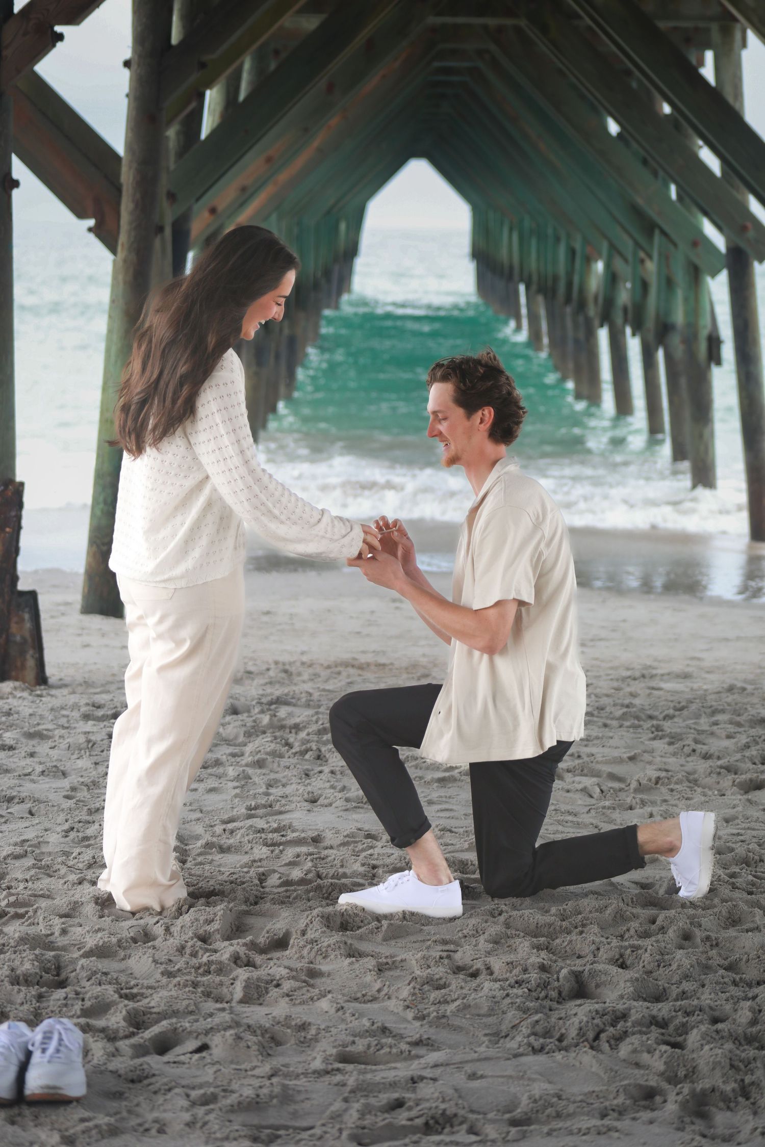 Man proposes to woman on one knee on sandy beach under wooden pier with turquoise ocean waves in background.