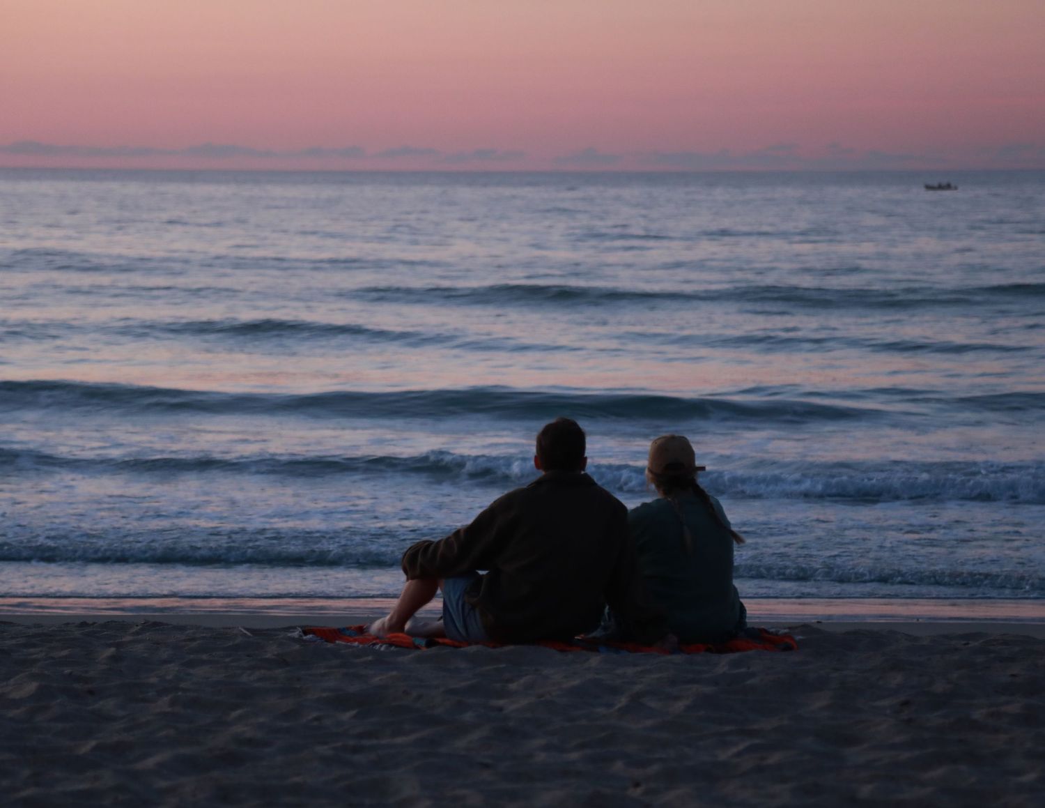 Two people sitting together on a sandy beach at sunset, watching calm ocean waves under a pink and purple sky.