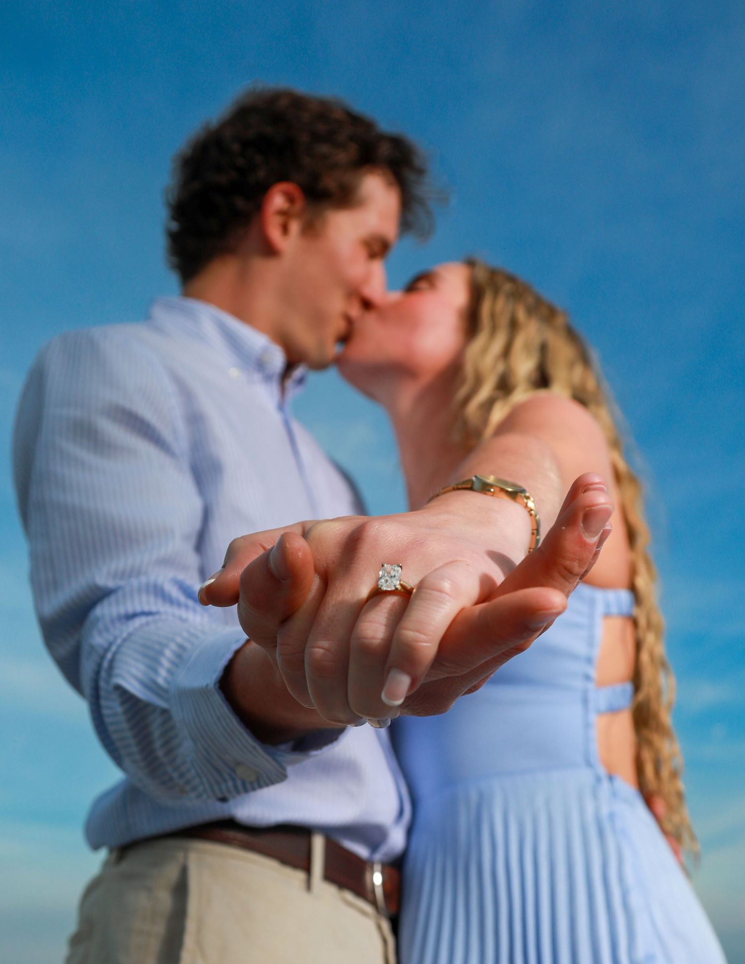 Couple kissing while showing off engagement ring with blue sky background.