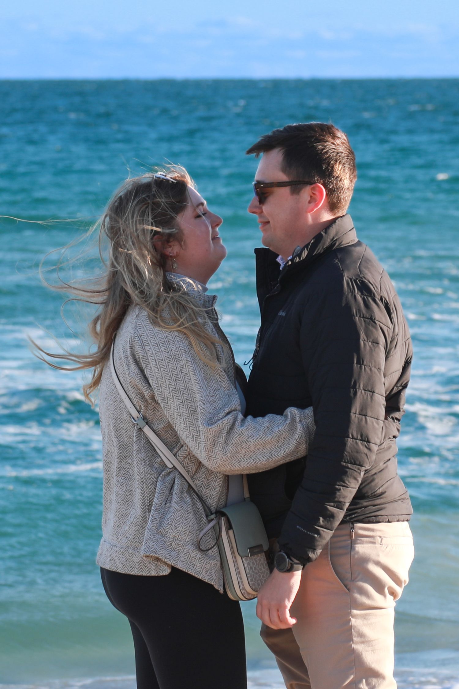 Couple embracing and gazing at each other on a windy beach, wearing jackets with turquoise ocean waves behind them.