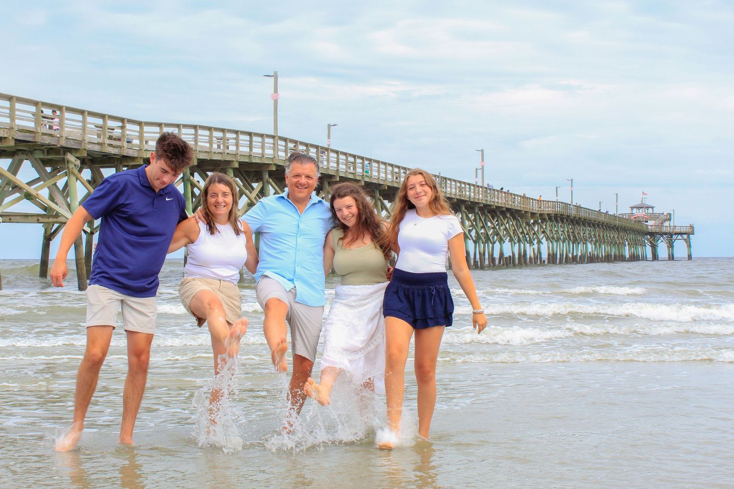 Happy family of five splashing in ocean waves near a pier on a sunny beach day.