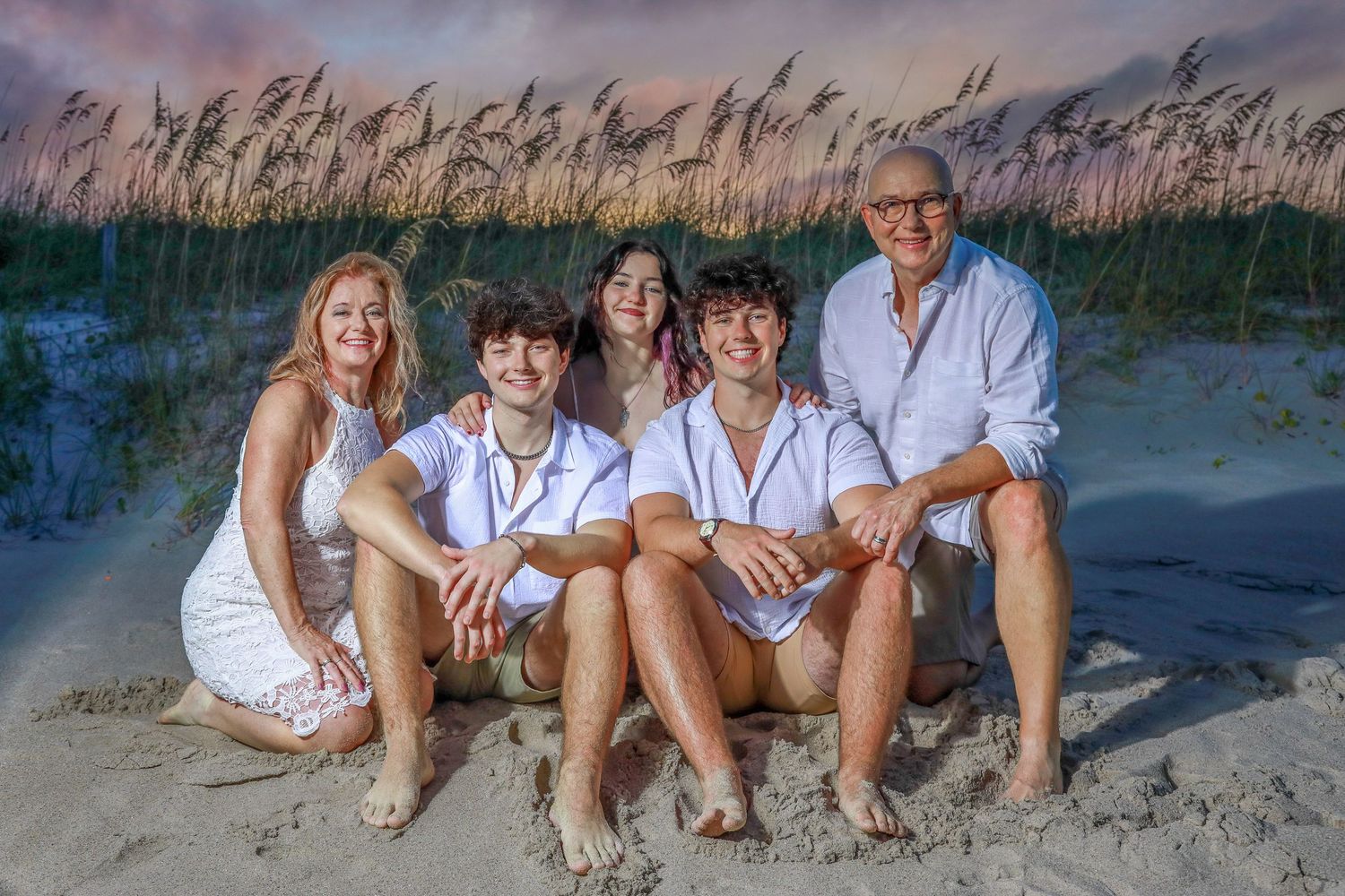 Family of five smiling together on a sandy beach at sunset, wearing coordinated white outfits with sea oats in background.