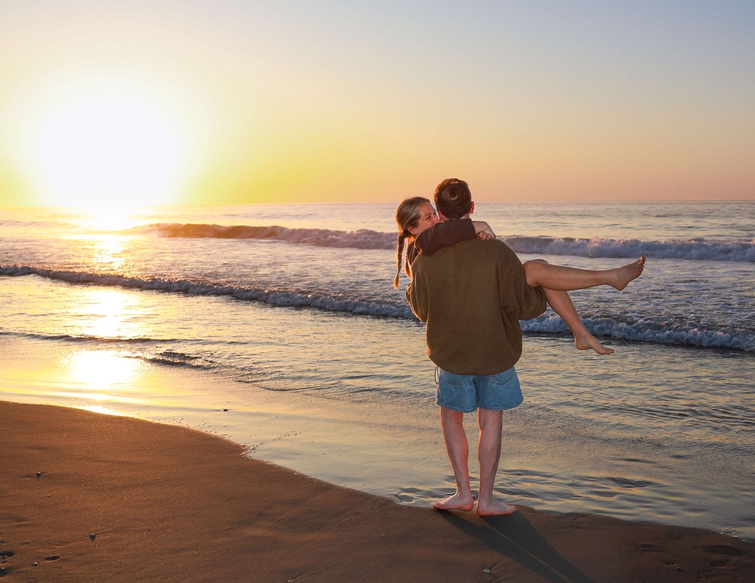 Couple embracing at sunset on a sandy beach, man carrying woman in his arms with ocean waves in the background.