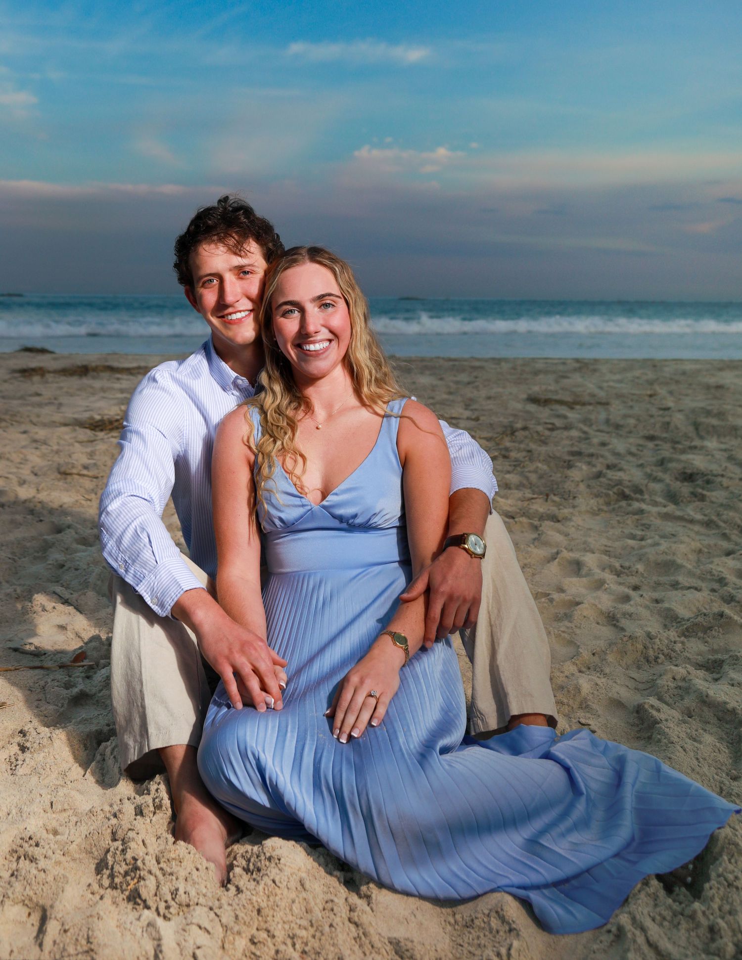 Happy couple sitting on sandy beach at sunset, woman in blue dress and man in white shirt smiling.