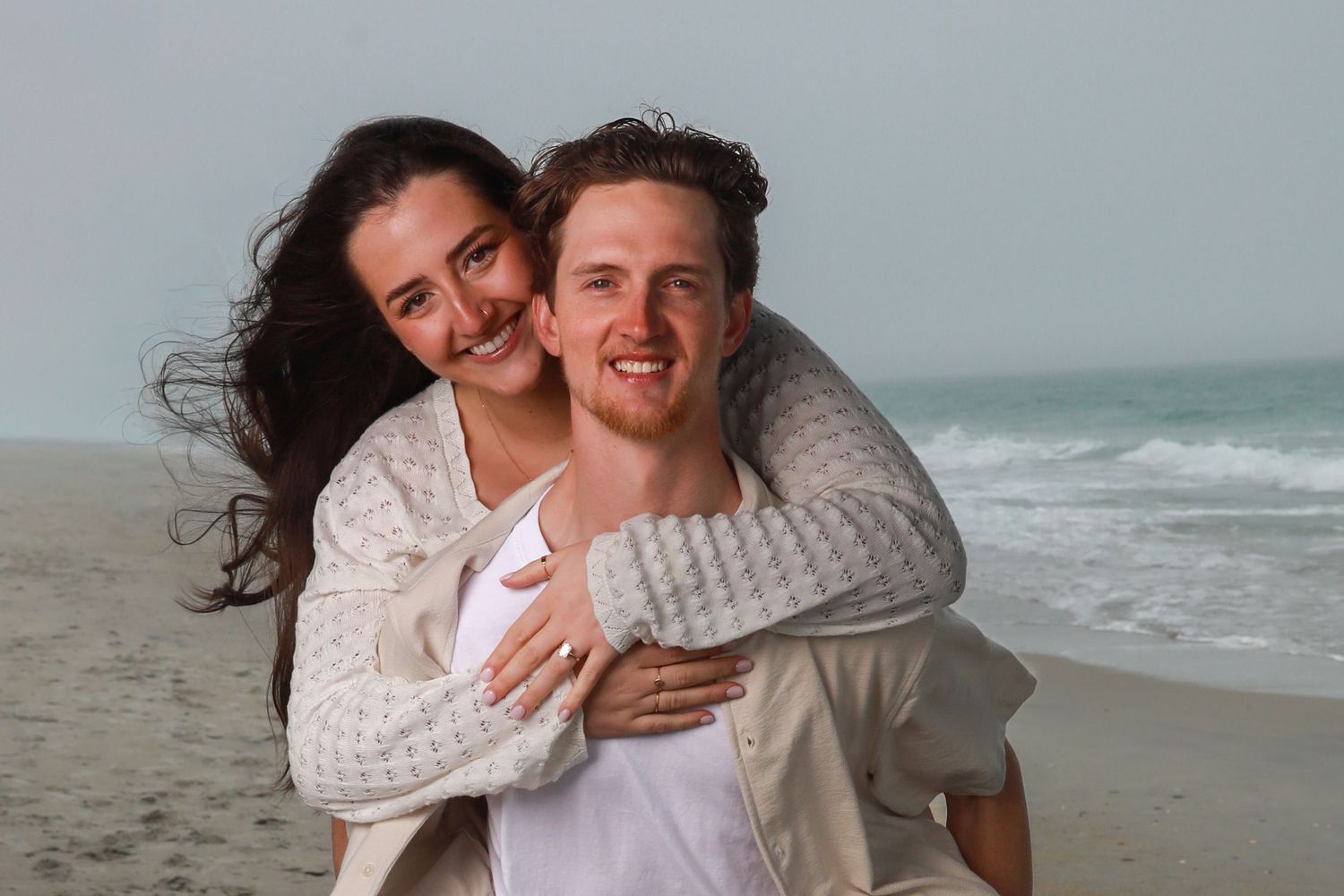 Happy couple embracing on a beach, woman piggyback on man, smiling with ocean waves in the background.