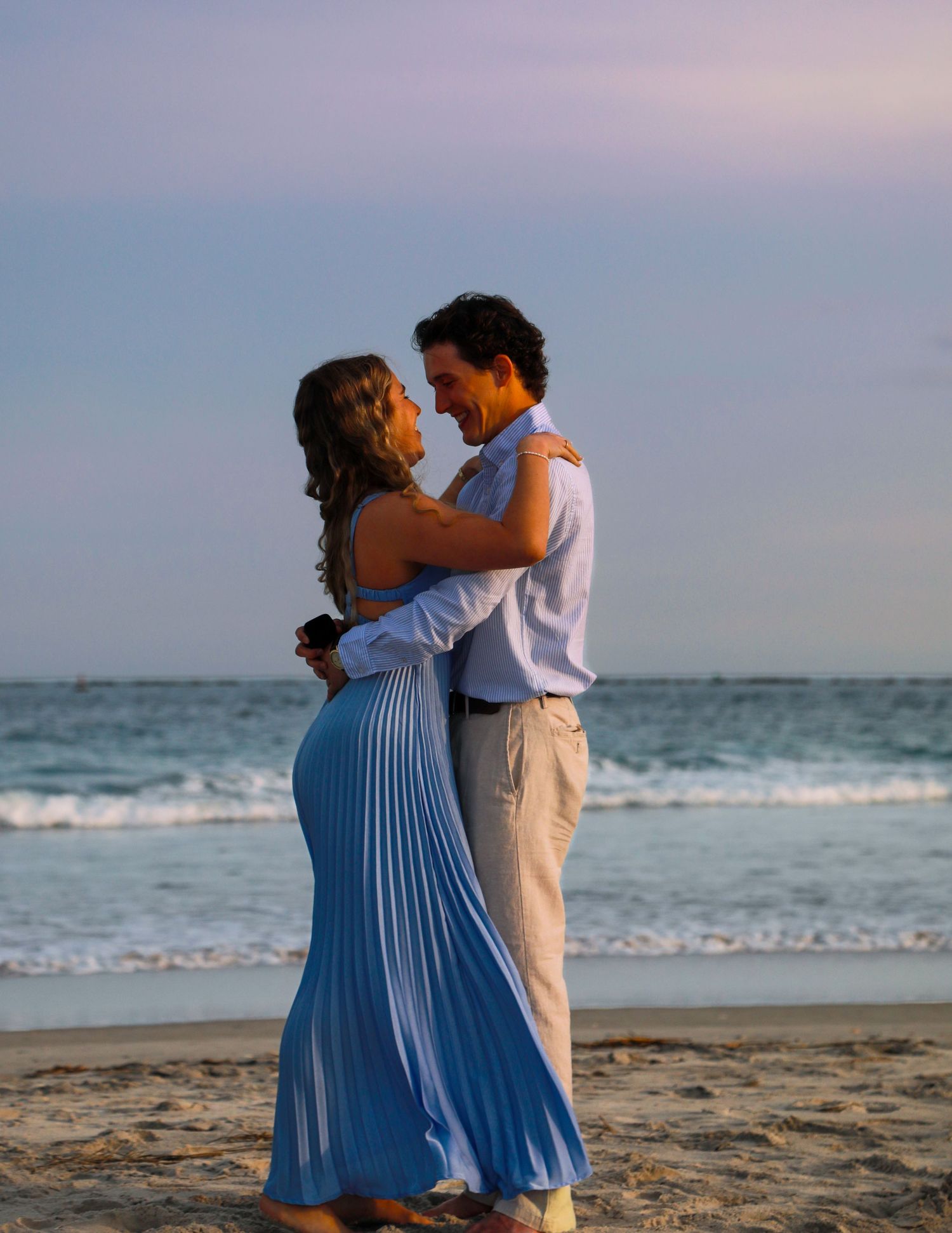 Couple embracing romantically on a beach at sunset, woman in blue pleated dress, man in white shirt and khaki pants.