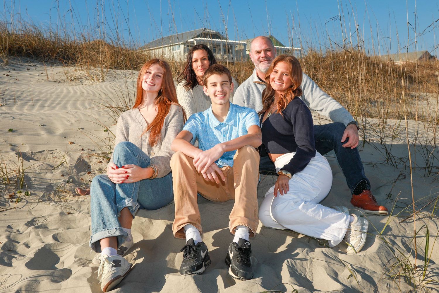 Family members sit together on a sandy beach with tall grass and dunes in the background during golden hour.