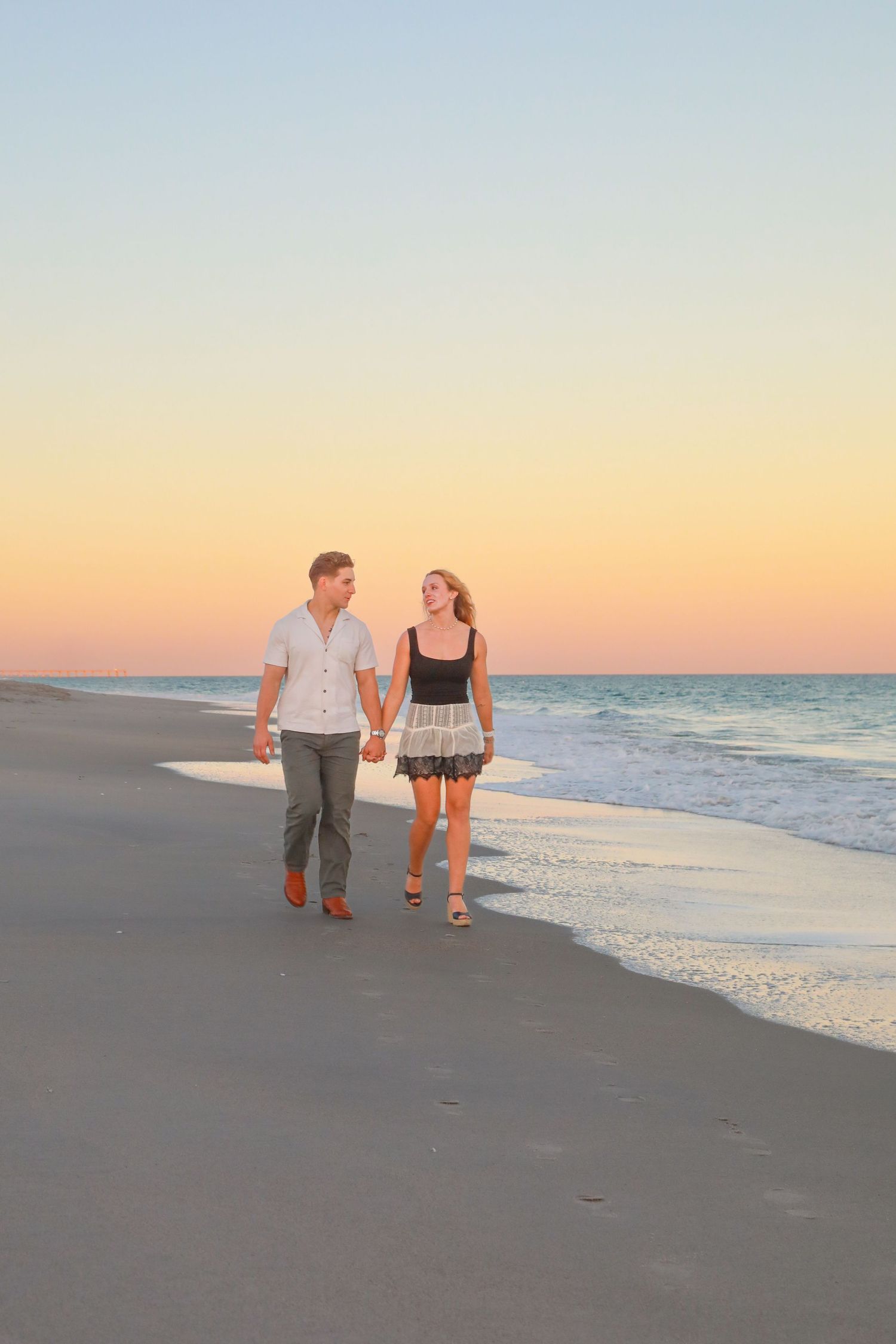 Couple holding hands walking on a sandy beach at sunset with ocean waves in the background.
