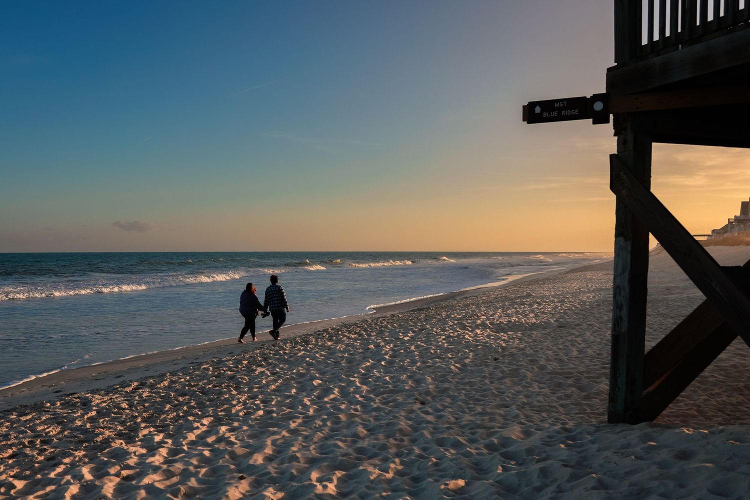 Two silhouetted figures walk along a sandy beach at sunset beside a lifeguard tower with golden light reflecting on the waves.