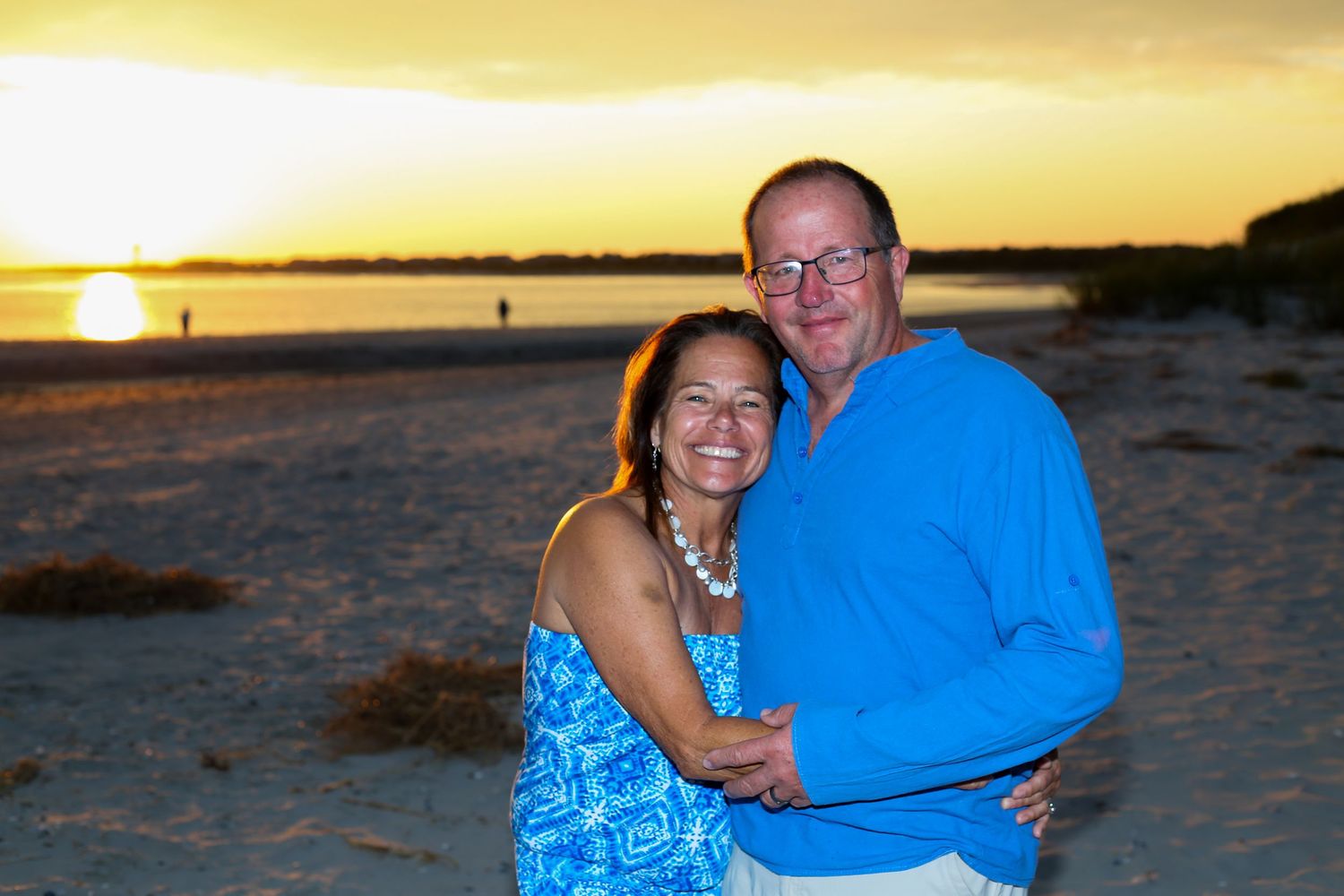 Couple smiling and embracing on a sandy beach at sunset with golden sky and ocean in the background.