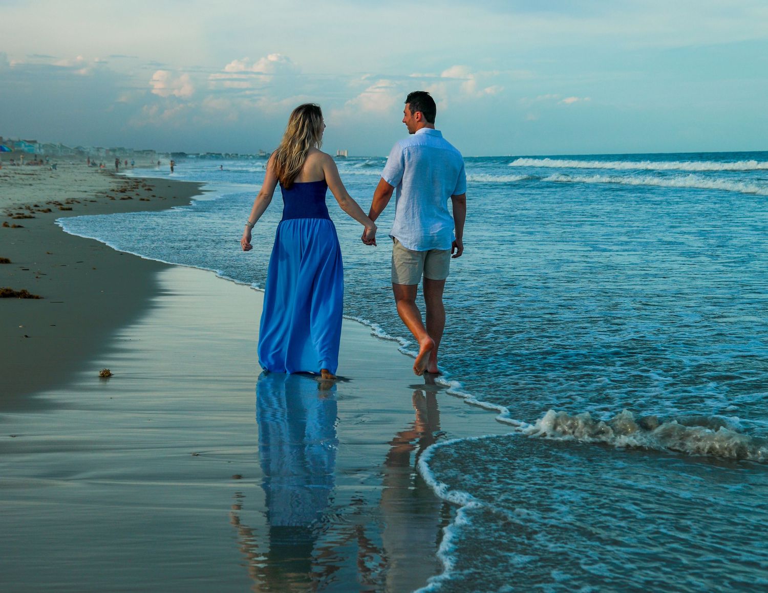 Couple holding hands walking along a serene beach shoreline at dusk, woman in blue maxi dress.