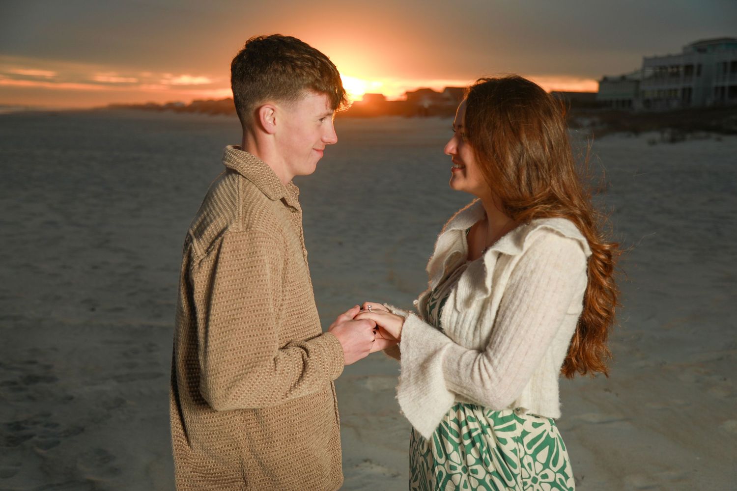 Couple sharing a romantic beach proposal at sunset, man placing ring on woman's finger by the ocean.
