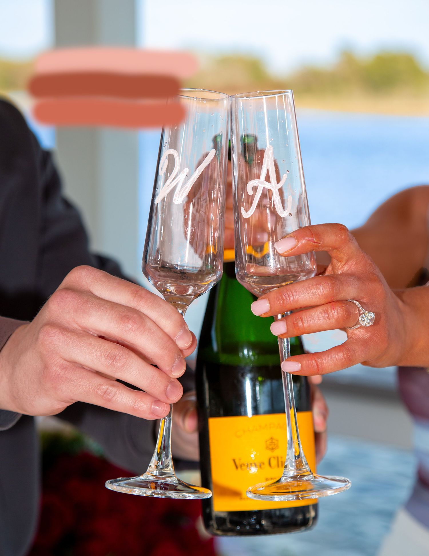 Two monogrammed champagne flutes toasting over a Veuve Clicquot bottle, with an engagement ring visible on one hand.
