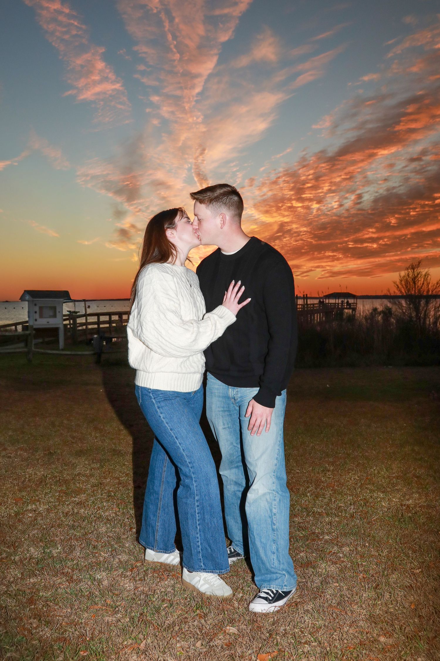A couple embraces during a stunning sunset with vibrant orange and pink clouds in a rural field setting.