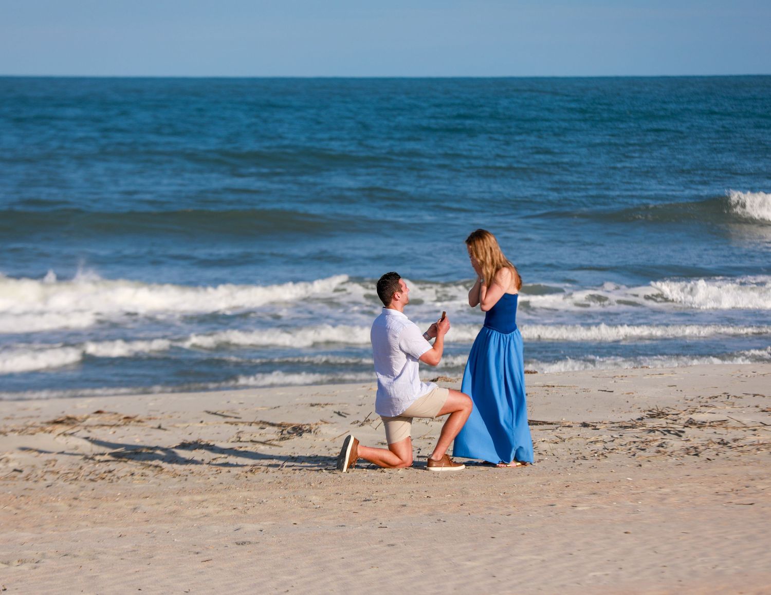 Man proposing to woman on a sandy beach with ocean waves, she wears a blue dress in this romantic moment.