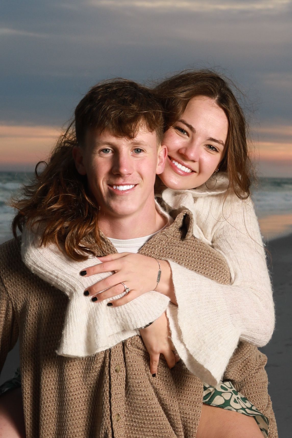Happy young couple embracing on a beach at sunset, smiling warmly in cozy sweaters with ocean waves behind them.