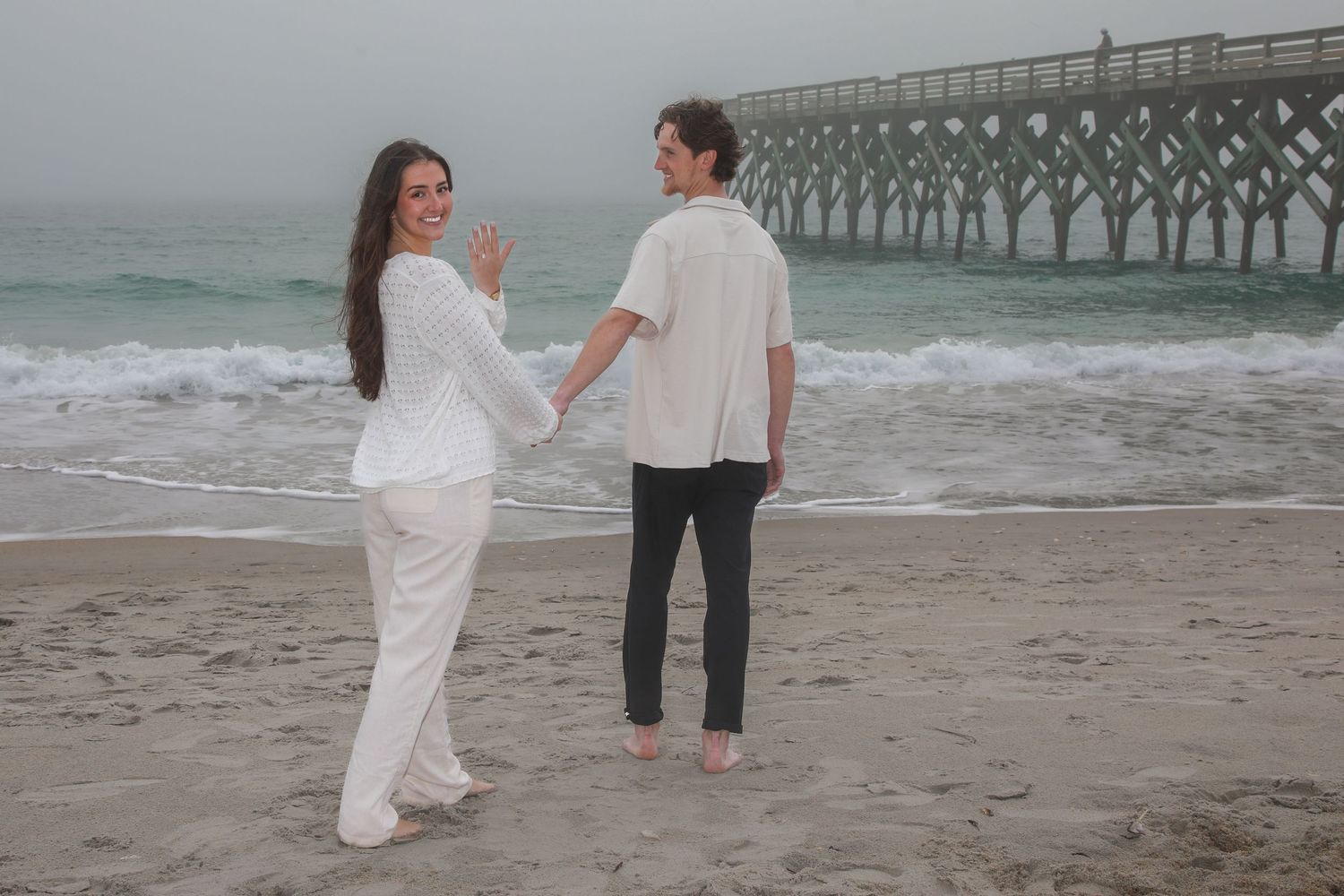 Couple holding hands on a misty beach near a pier, woman in white pantsuit showing ring, man in white shirt.