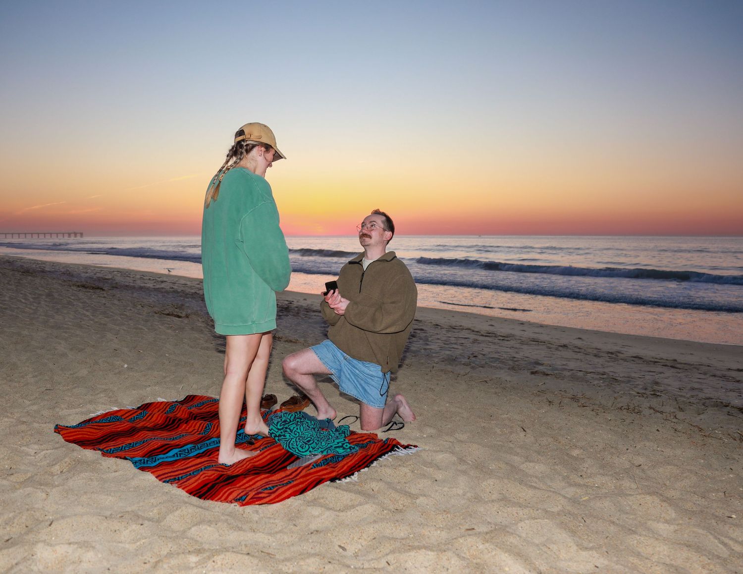Man proposing to woman on a colorful blanket at a beach during a stunning sunset.