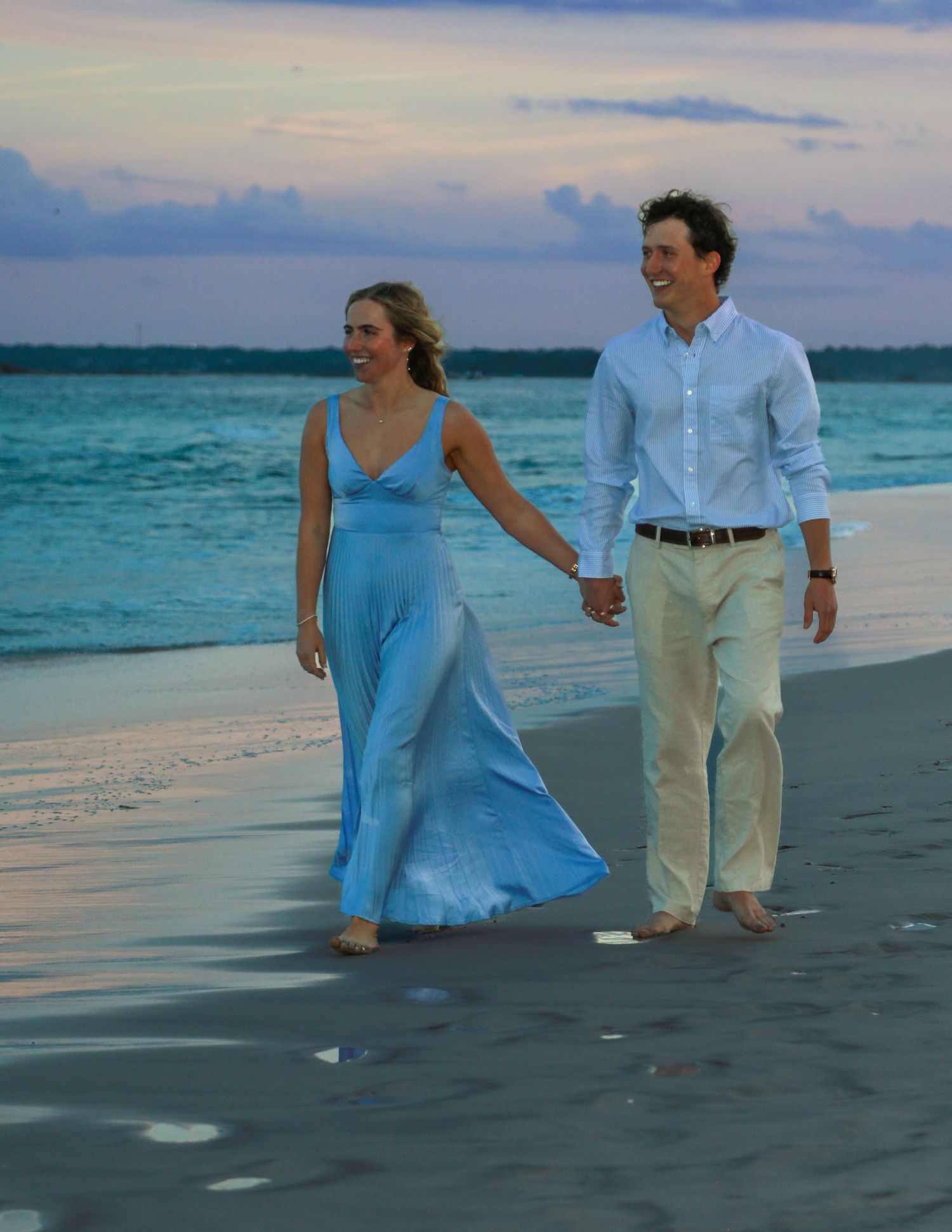 Couple holding hands walking on a beach at sunset, woman in blue maxi dress, man in light blue shirt and khaki pants.