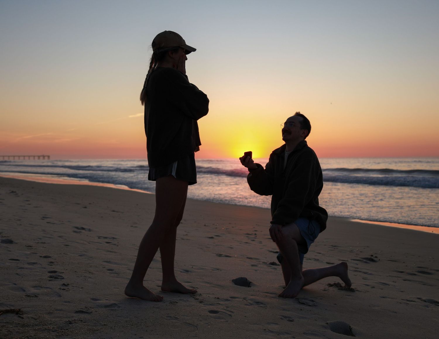 Silhouette of a man proposing on one knee to a woman on a beach at sunset with golden sky.