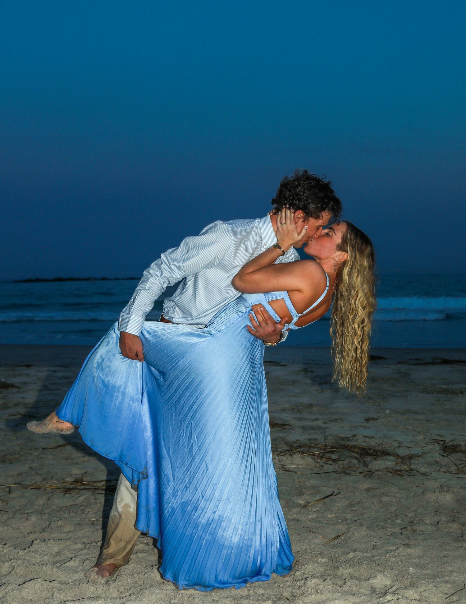 Couple sharing a romantic dip kiss on a beach at dusk, woman in blue gown, man in white shirt.