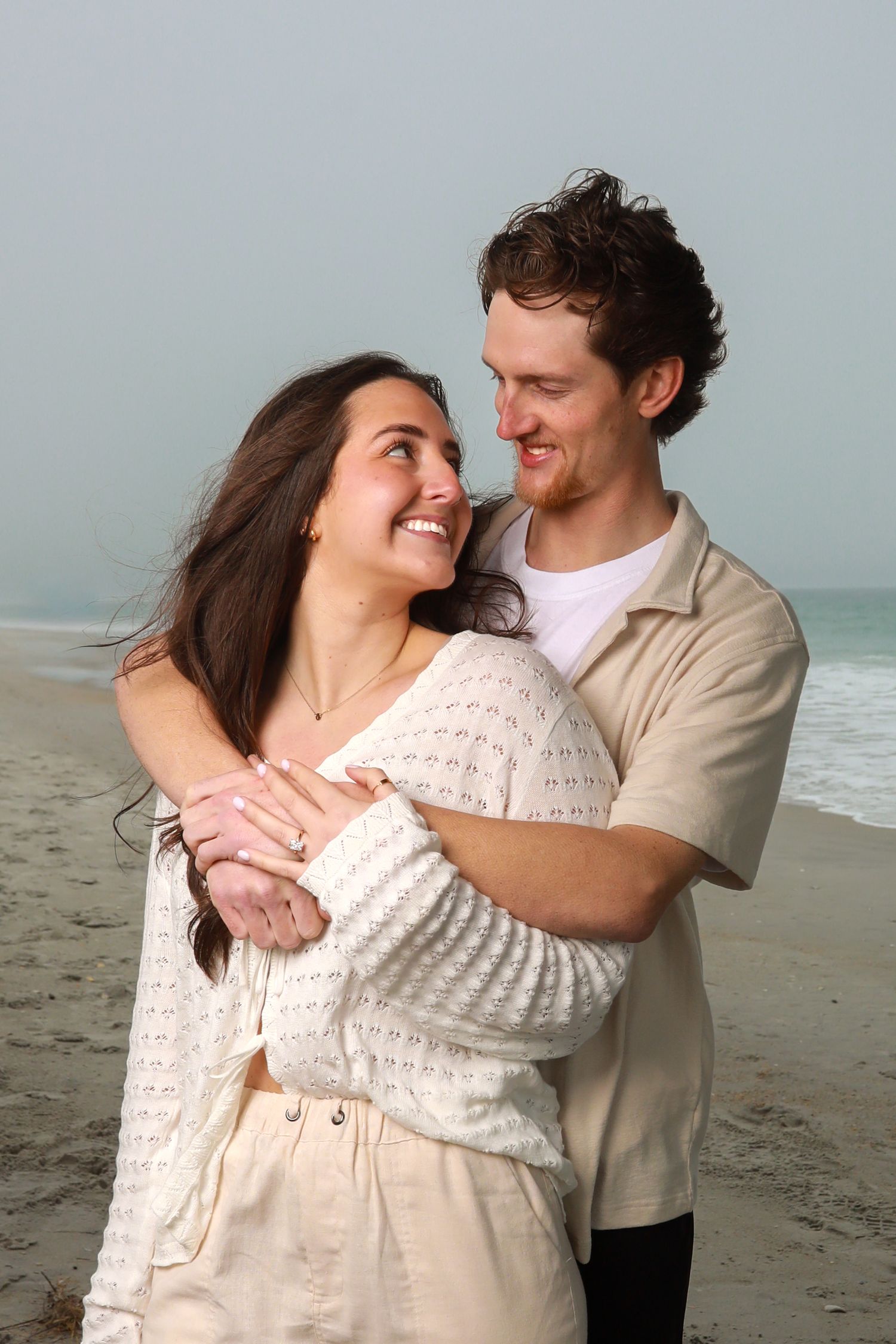 Happy couple embracing on a beach, smiling at each other in matching beige outfits with ocean waves in the background.