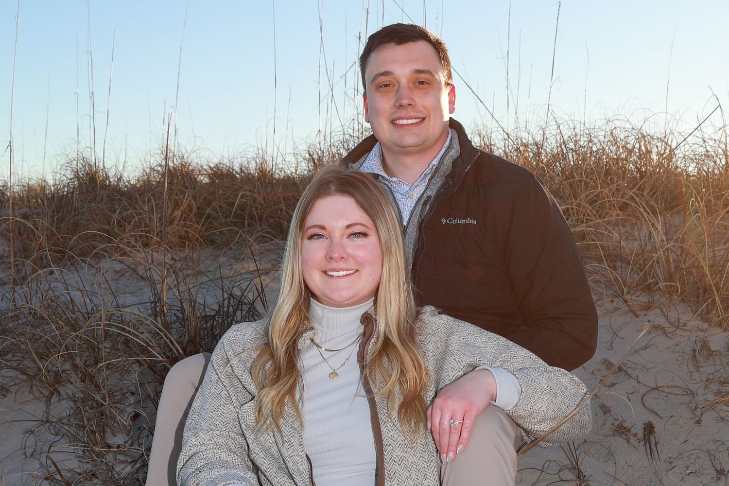 A smiling couple poses together on a sandy beach with dune grass in the background on a sunny day.