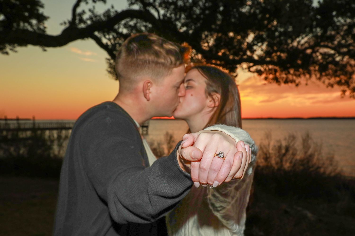 A romantic couple embraces and kisses during a beautiful sunset by a lake with golden light illuminating the scene.